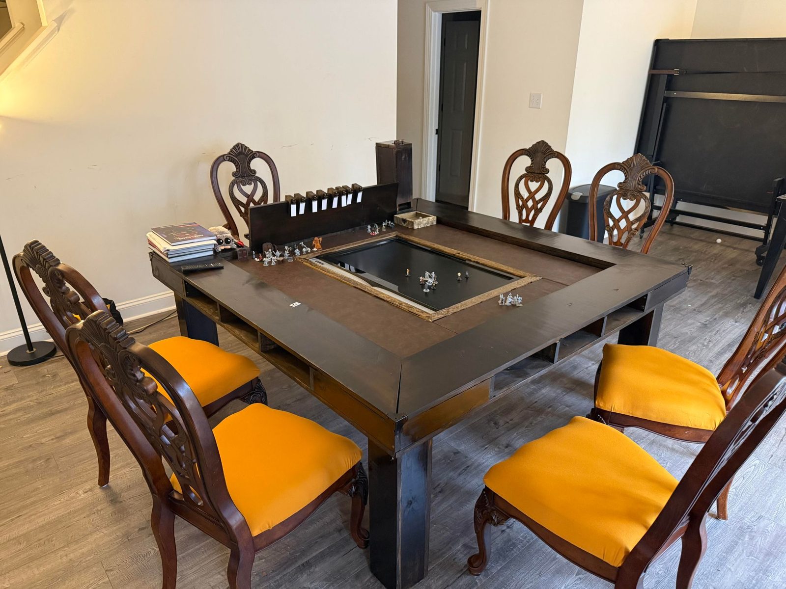 A dark wood gaming table with a recessed center screen for displaying maps, miniatures set out mid-session, and six chairs with gold cushions around it.